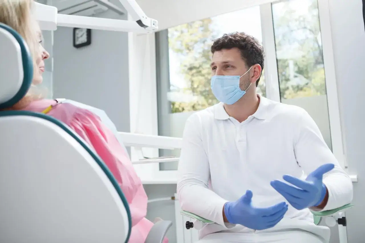 Dentysta w masce omawia antybiotyki w stomatologii z pacjentką siedzącą na fotelu.