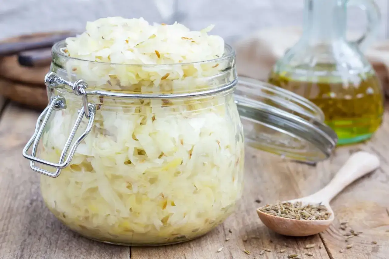 Ein Glas Sauerkraut, das für eine gute Verdauung bekannt ist, steht auf einem Holztisch. Daneben eine Karaffe Öl und ein Löffel mit Kümmelsamen.