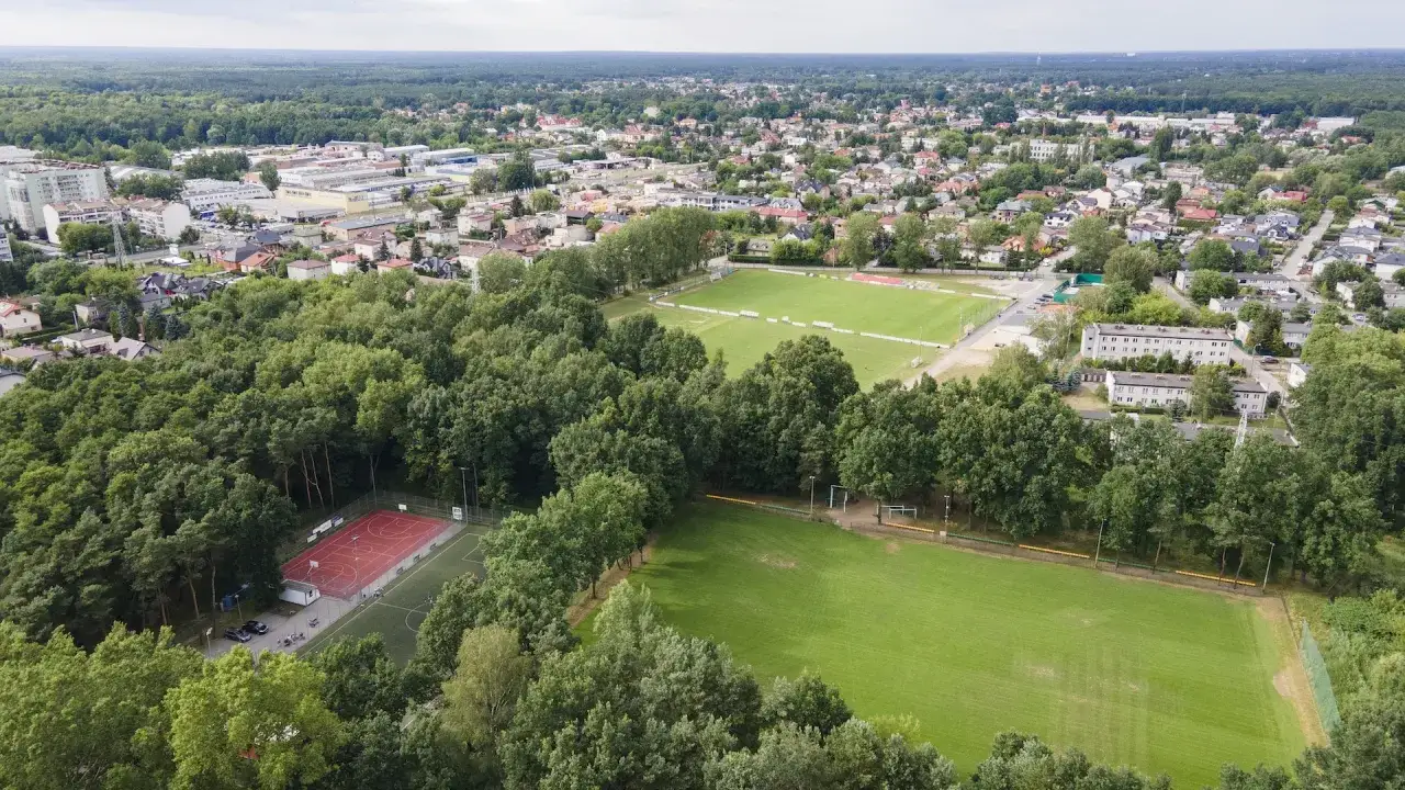 Widok z lotu ptaka na miasto z boiskami sportowymi, kortem tenisowym i terenami zielonymi. W tle widać zabudowania i las. Idealne miejsce dla miłośników sportu i aktywności na świeżym powietrzu, gdzie różne **marki sportowe** mają swoje zaplecze.