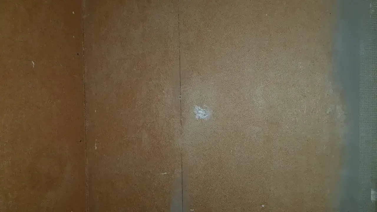 En vägg av masonit som håller på att målas. En del är täckt av ljusgrå färg, medan en annan del fortfarande är brun.