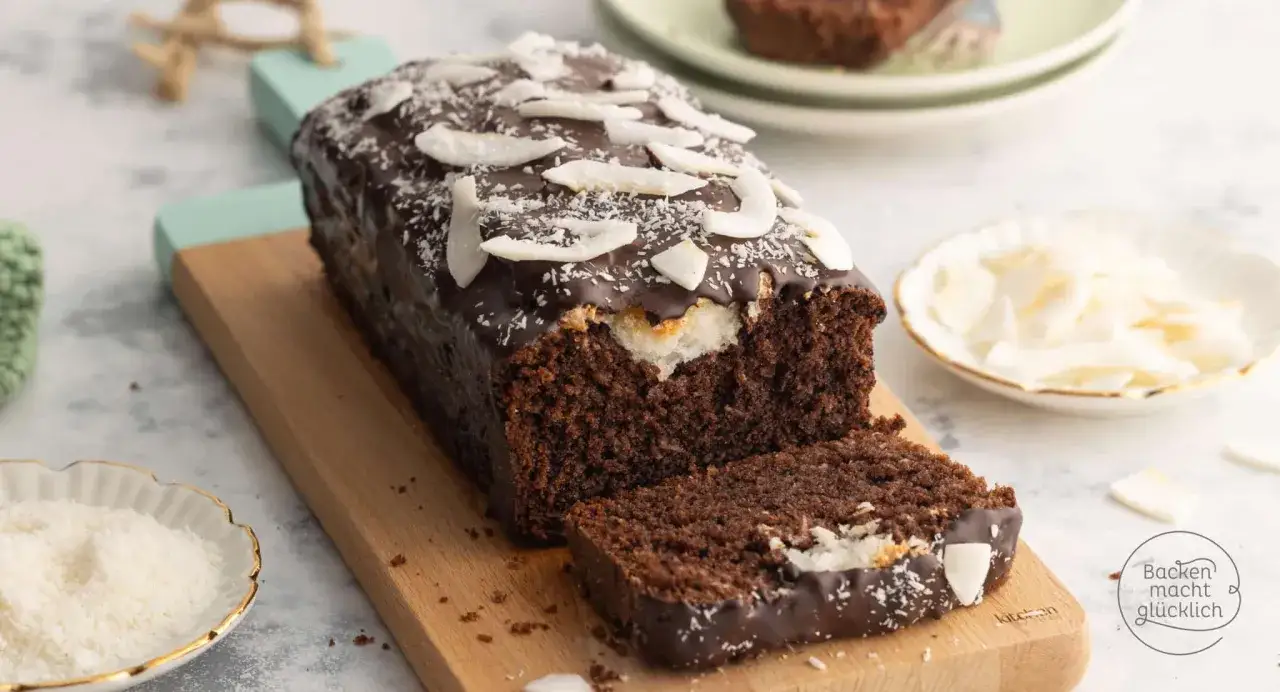 Schoko Kokos Kuchen mit dunkler Glasur und Kokosraspeln, ein St&uuml;ck angeschnitten, auf einem Holzbrett.