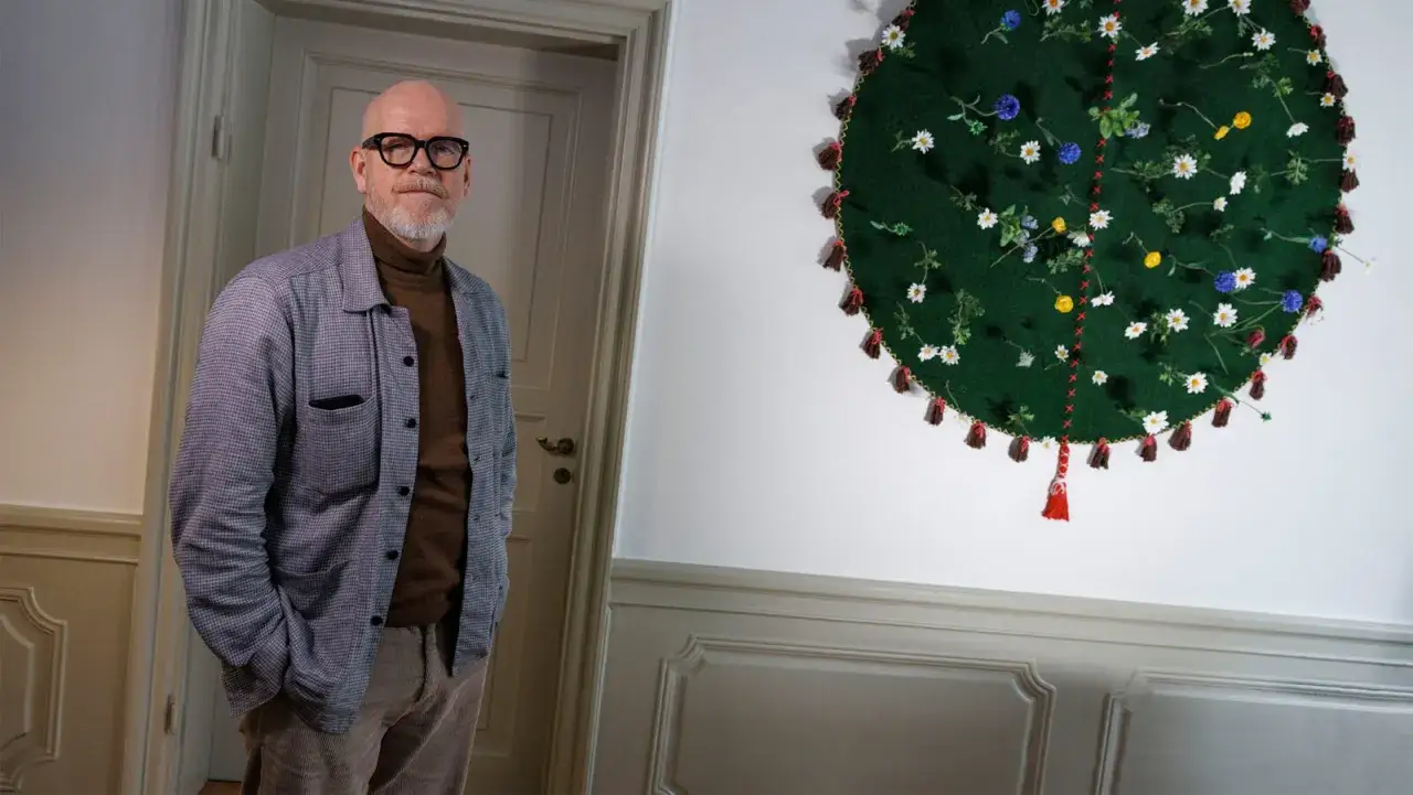Jonas Axelsson, en man med glasögon och skägg, står bredvid ett runt, grönt konstverk med blommor och tofsar.