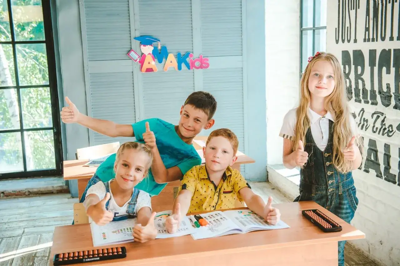 Dzieci z kciukami w g&oacute;rze, uczące się arytmetyki. Na stole liczydła i zeszyty.