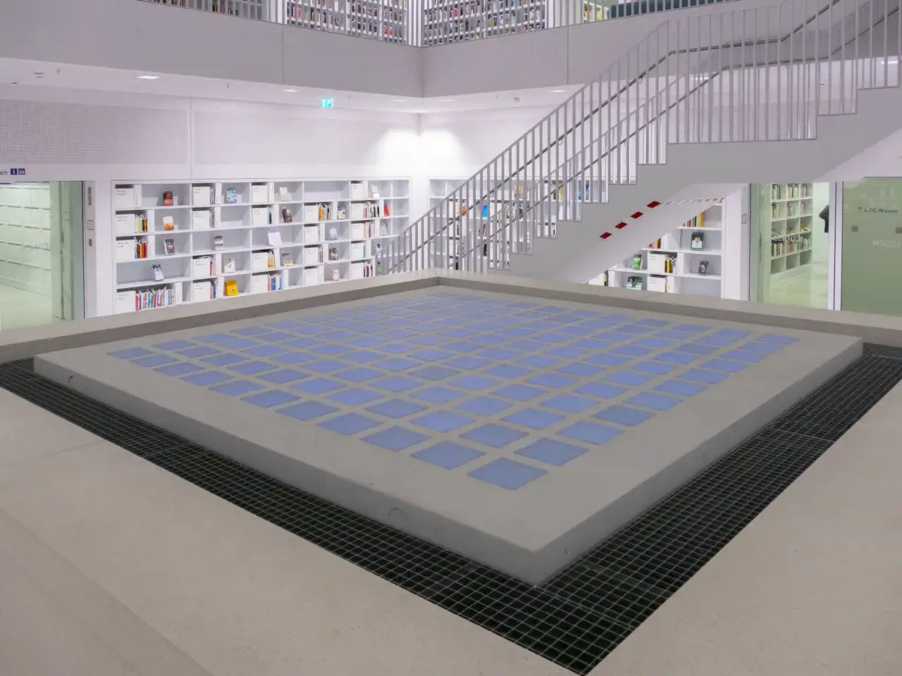 Moderne Stuttgart Bibliothek mit wei&szlig;en Regalen voller B&uuml;cher, einer Treppe und einem zentralen Platz mit blauen Quadraten.