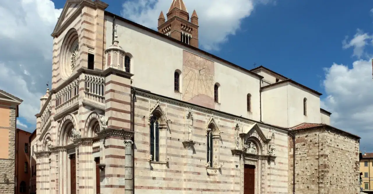 Il duomo di Grosseto, con la sua facciata a strisce bianche e rosa, torre campanaria e un grande quadrante solare sulla parete laterale, si staglia contro un cielo azzurro con nuvole bianche.