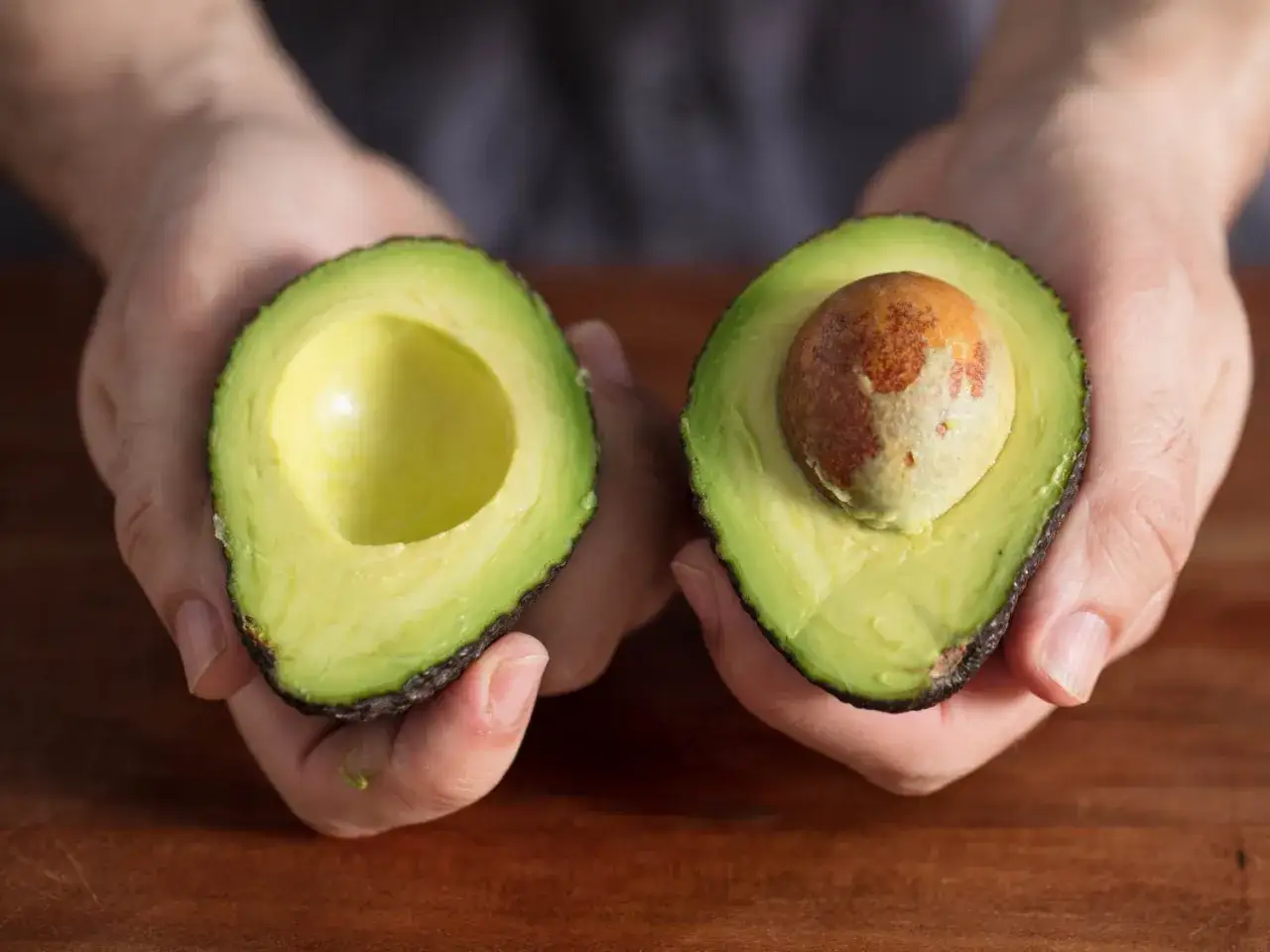 Zwei Hälften einer Avocado in Händen. Eine ist leer, die andere mit Kern. Geduld ist gefragt, um die Avocado reifen lassen.