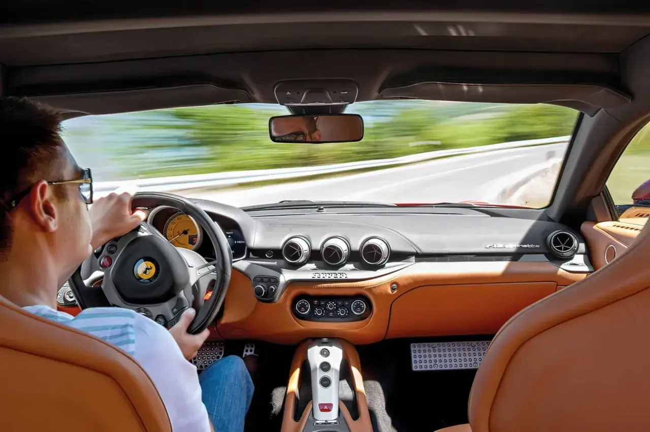 Wnętrze Ferrari F12 Berlinetta. Kierowca w okularach prowadzi auto po krętej drodze, otoczony luksusową, skórzaną tapicerką.