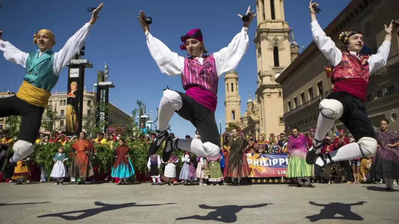 Jóvenes bailan jotas del Pilar con castañuelas en alto, saltando con energía ante una multitud y edificios históricos.