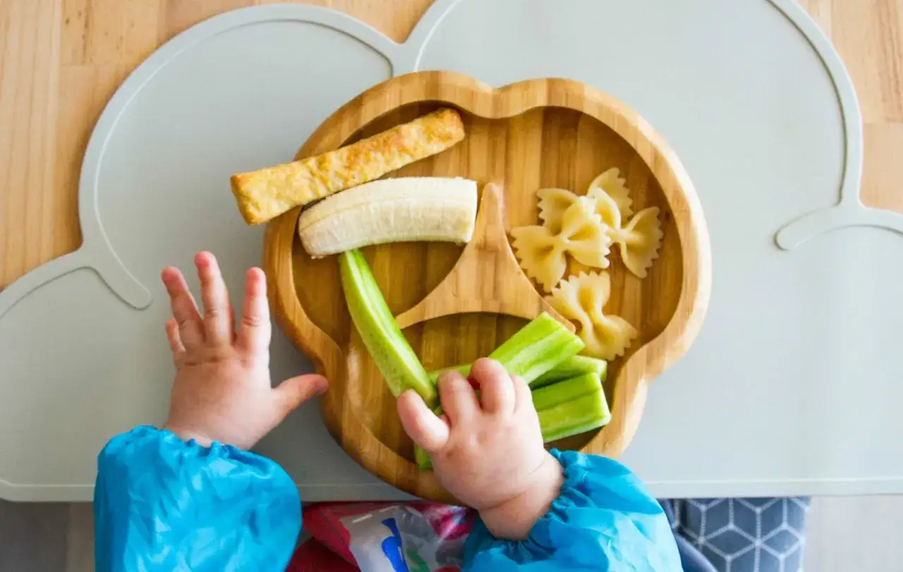 Babyhände greifen nach Nudeln, Banane und Gurke auf einem Holzbrettchen – ein bunter beikostplan baby.