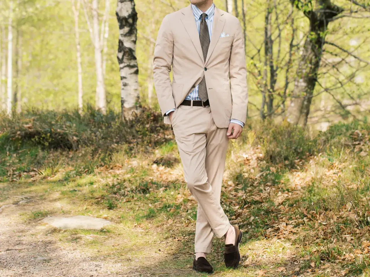 Man i ljusbeige kostym med slips och mockasiner, st&aring;ende i en skogsmilj&ouml;.