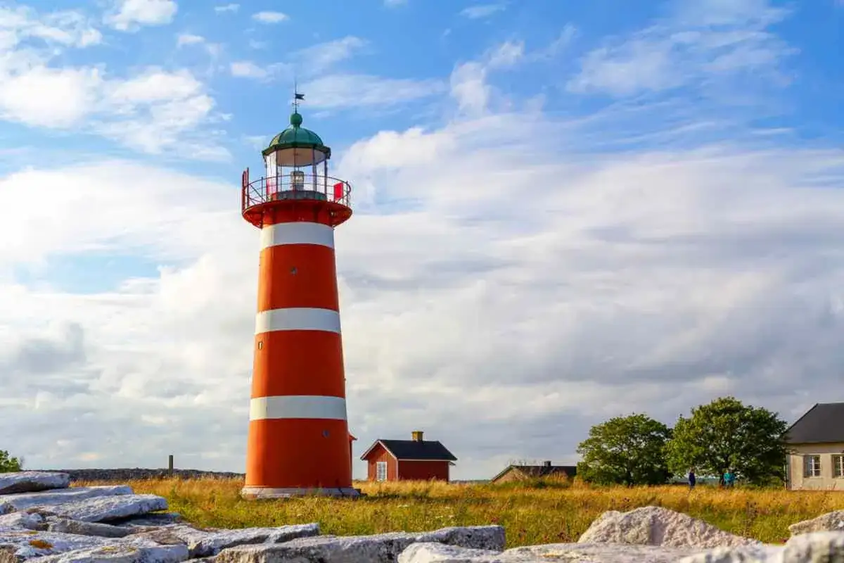 Rött och vitt fyr på Gotland, med små röda hus och gröna träd i bakgrunden under en molnig himmel.