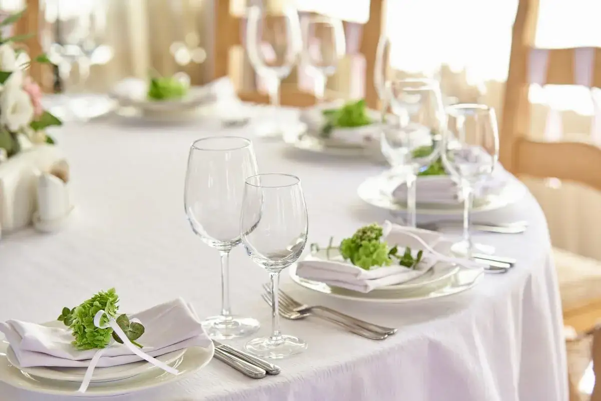 Comment dresser une table dans un restaurant : nappage blanc, verres à pied, assiettes, serviettes ornées de verdure et couverts.