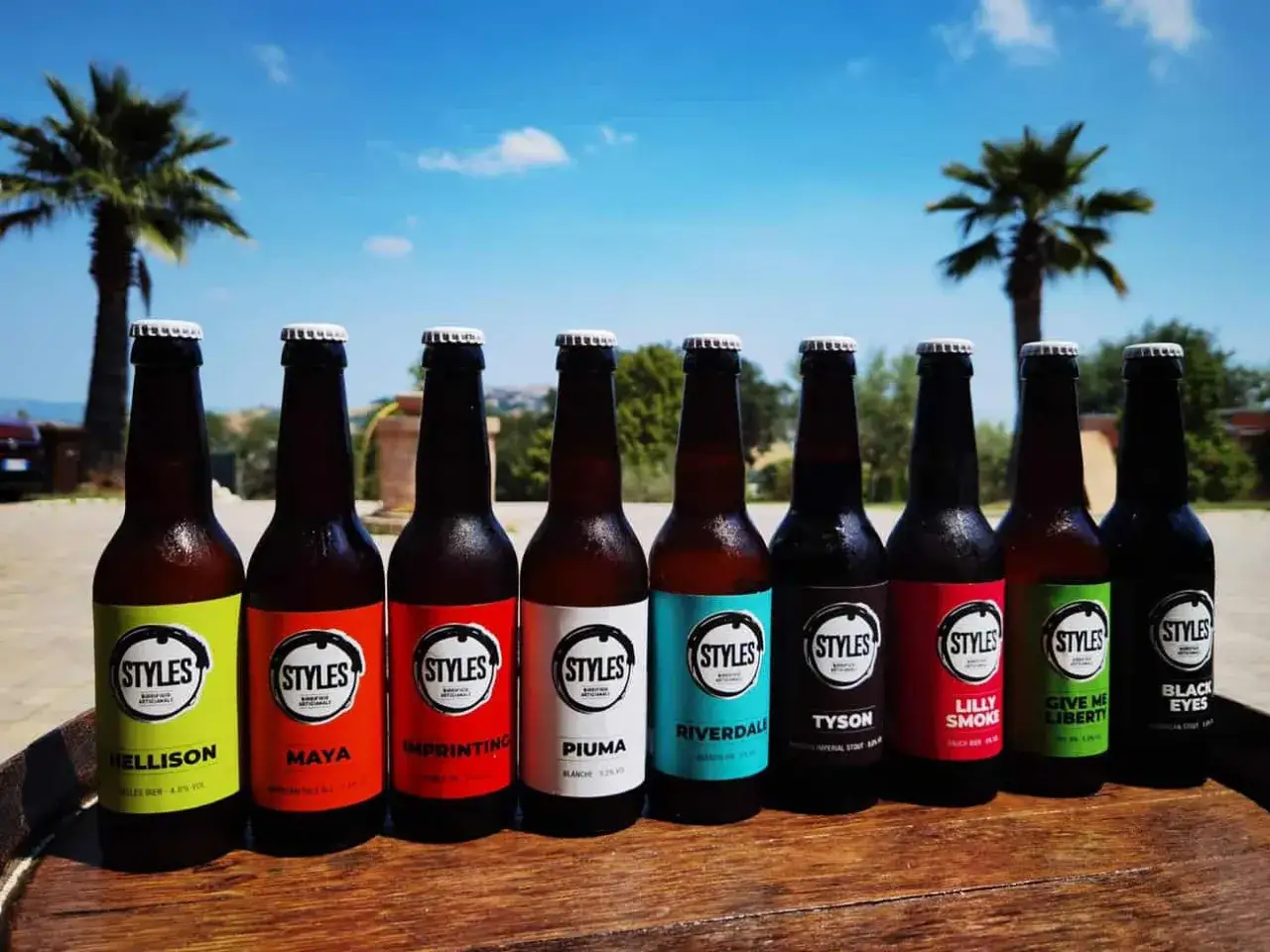 Otto bottiglie di birra Styles, tra cui la Hellison, Maya e Riverdale, pronte per essere gustate sotto il sole, con palme e cielo azzurro.