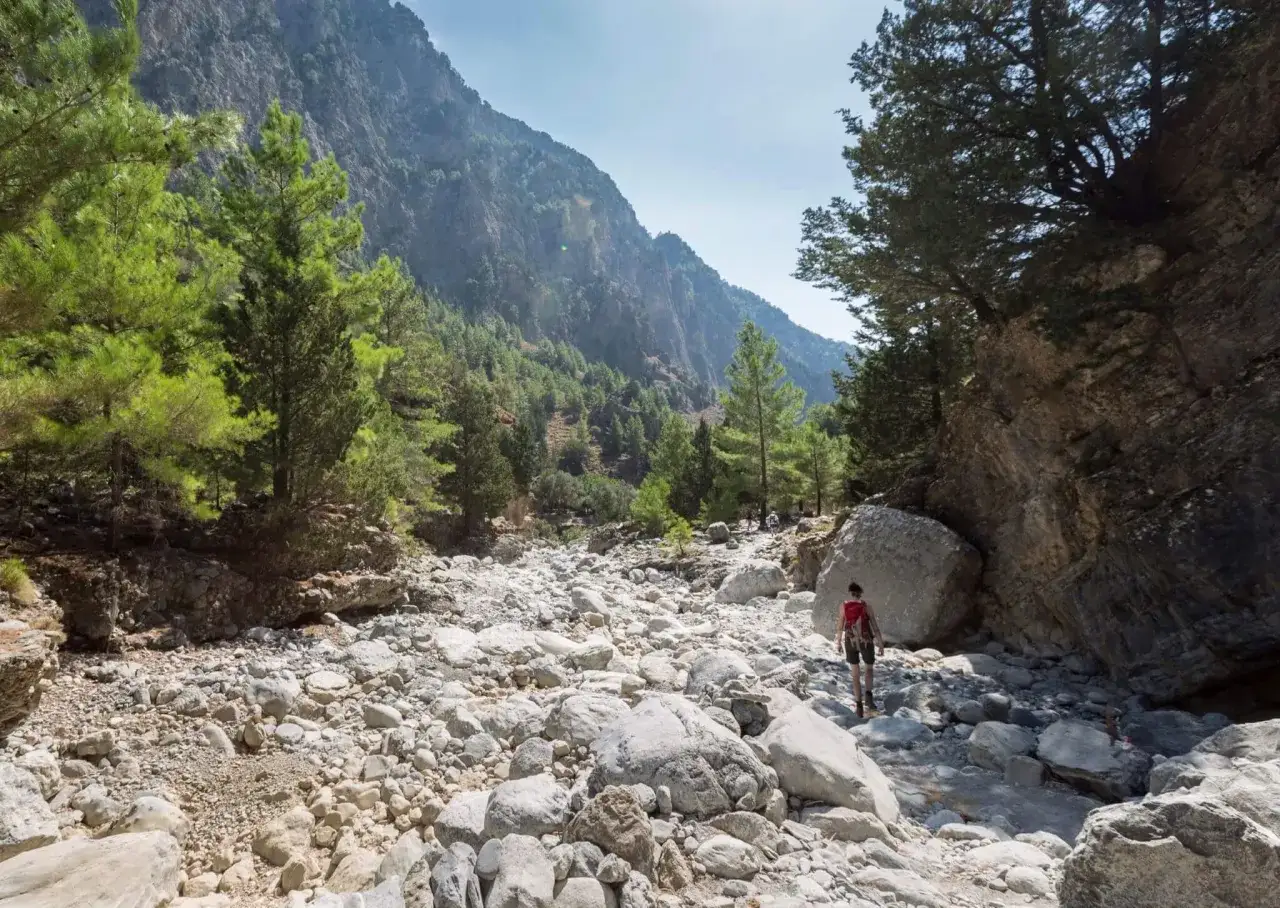 Wanderer mit rotem Rucksack durchquert die steinige Samaria Schlucht, umgeben von steilen Felsw&auml;nden und gr&uuml;nen B&auml;umen.