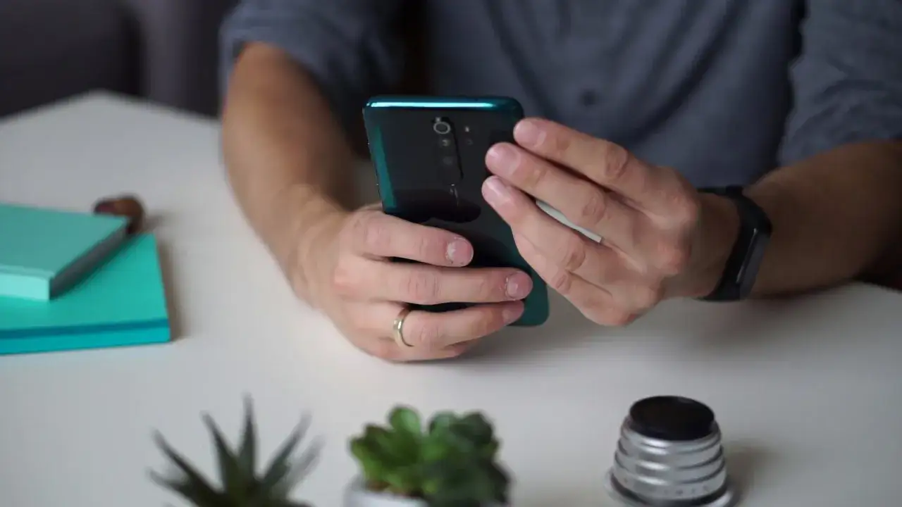 Mężczyzna trzyma Redmi Note 8 Pro, sprawdzając jego wymiary. Na stole leżą notatniki i obiektyw.