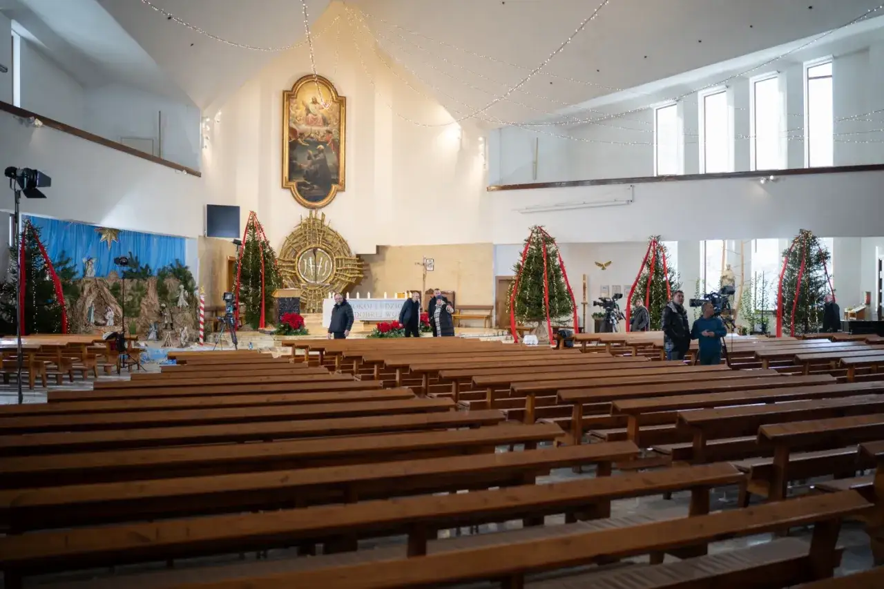 Wnętrze kościoła na Jastrzębiej Górze, udekorowane na Boże Narodzenie. Widać ławki, szopkę, choinki i ekipę telewizyjną.