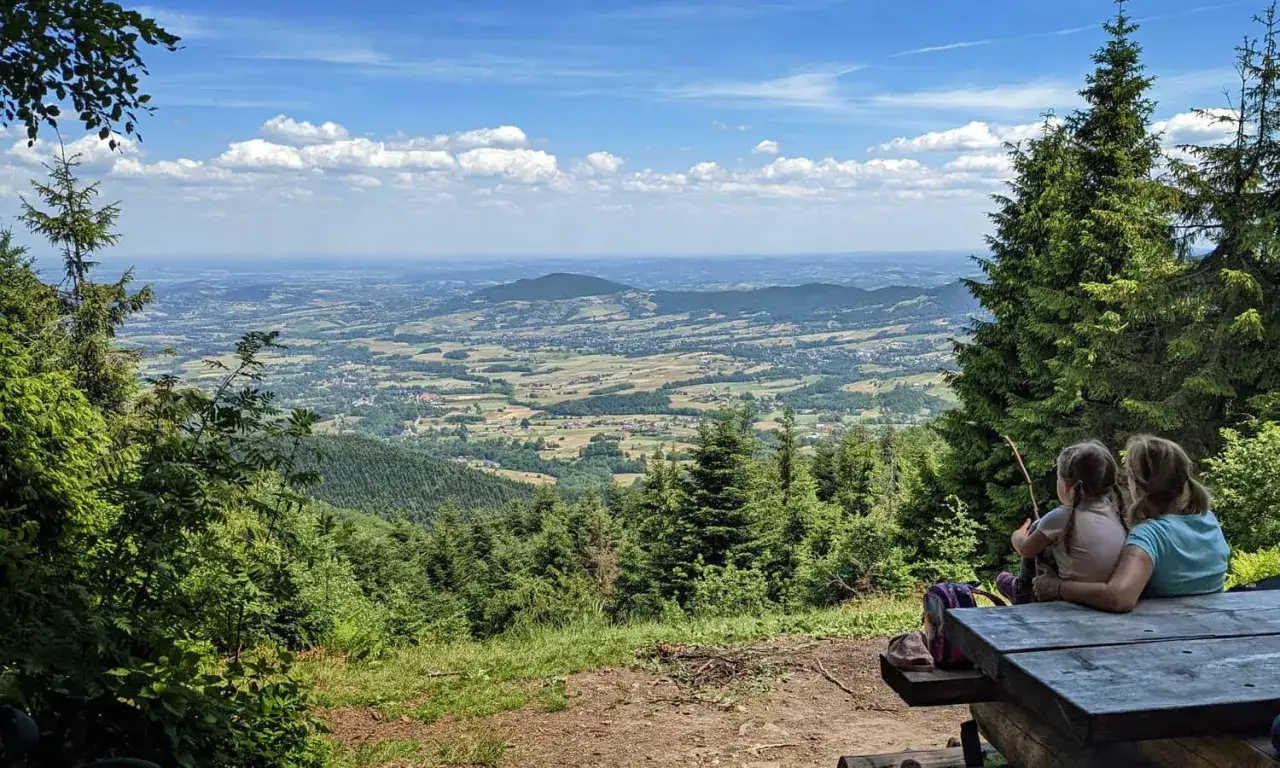 Dwie osoby siedzą przy drewnianym stole, podziwiając rozległą panoramę **Beskidu Makowskiego**. W oddali widać liczne **szczyty** i doliny.