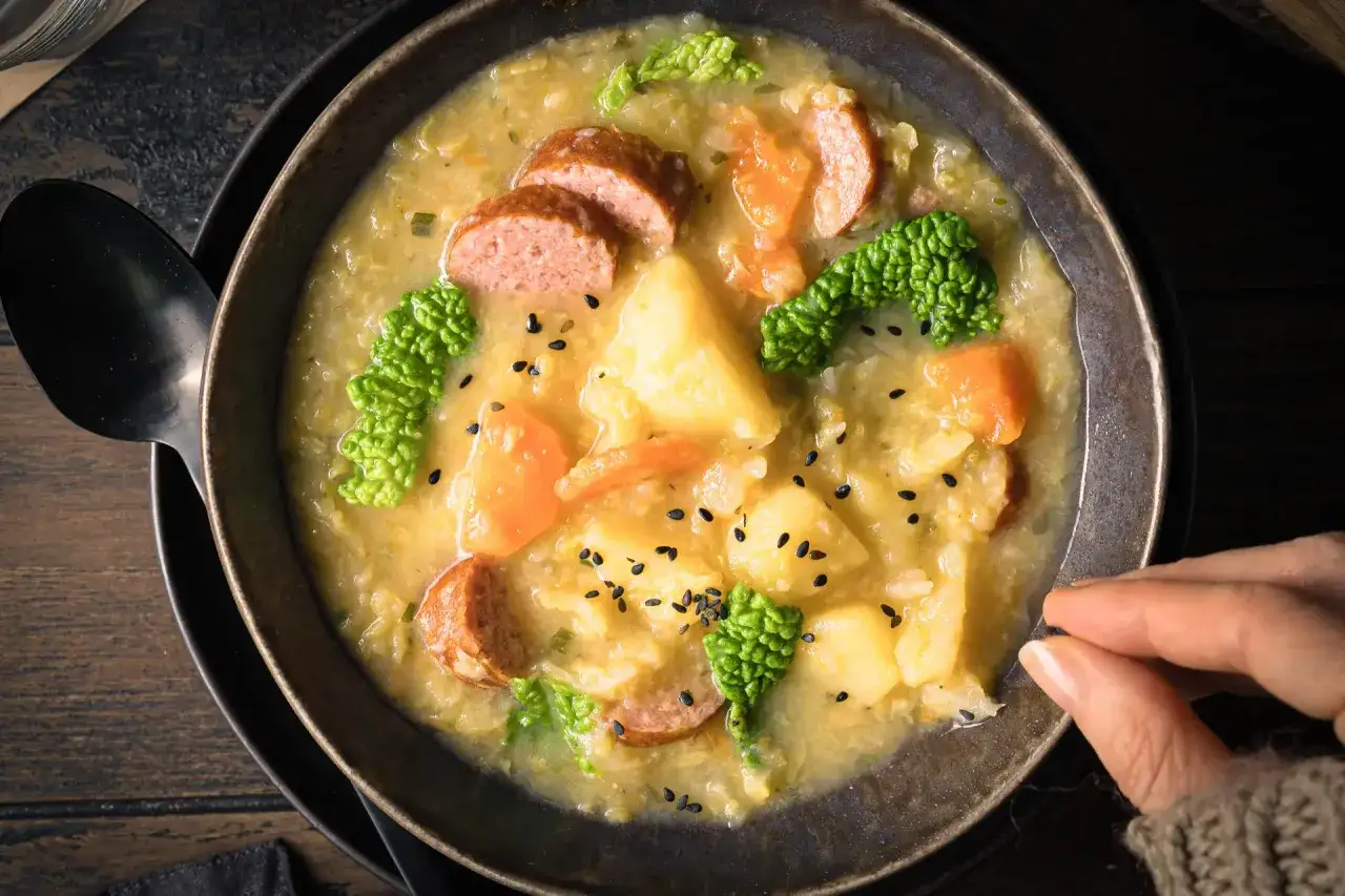 Herzhafte Suppe mit Wirsing, Kartoffeln, Karotten und Wurstscheiben, garniert mit schwarzen Sesamkörnern.
