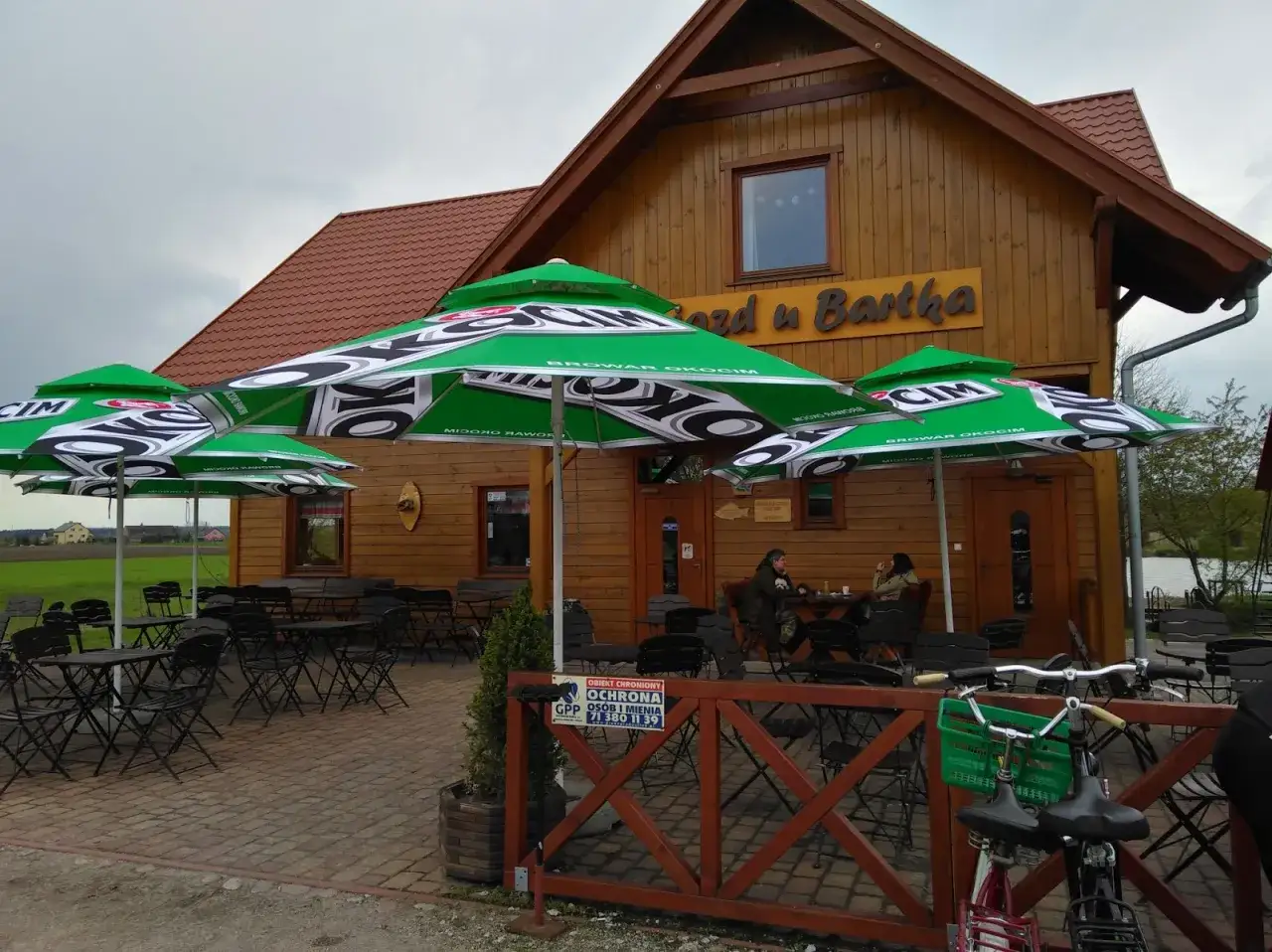 Zajazd u Bartka z zielonymi parasolami i drewnianym budynkiem. Dwie osoby siedzą przy stoliku na zewnątrz.