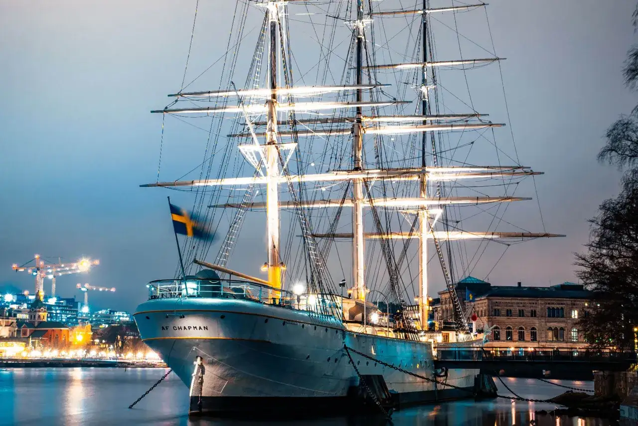 En st&aring;tlig skonare, AF Chapman, ligger vid kajen. En l&aring;ng rad av ljus lyser upp Skeppsholmen i bakgrunden.