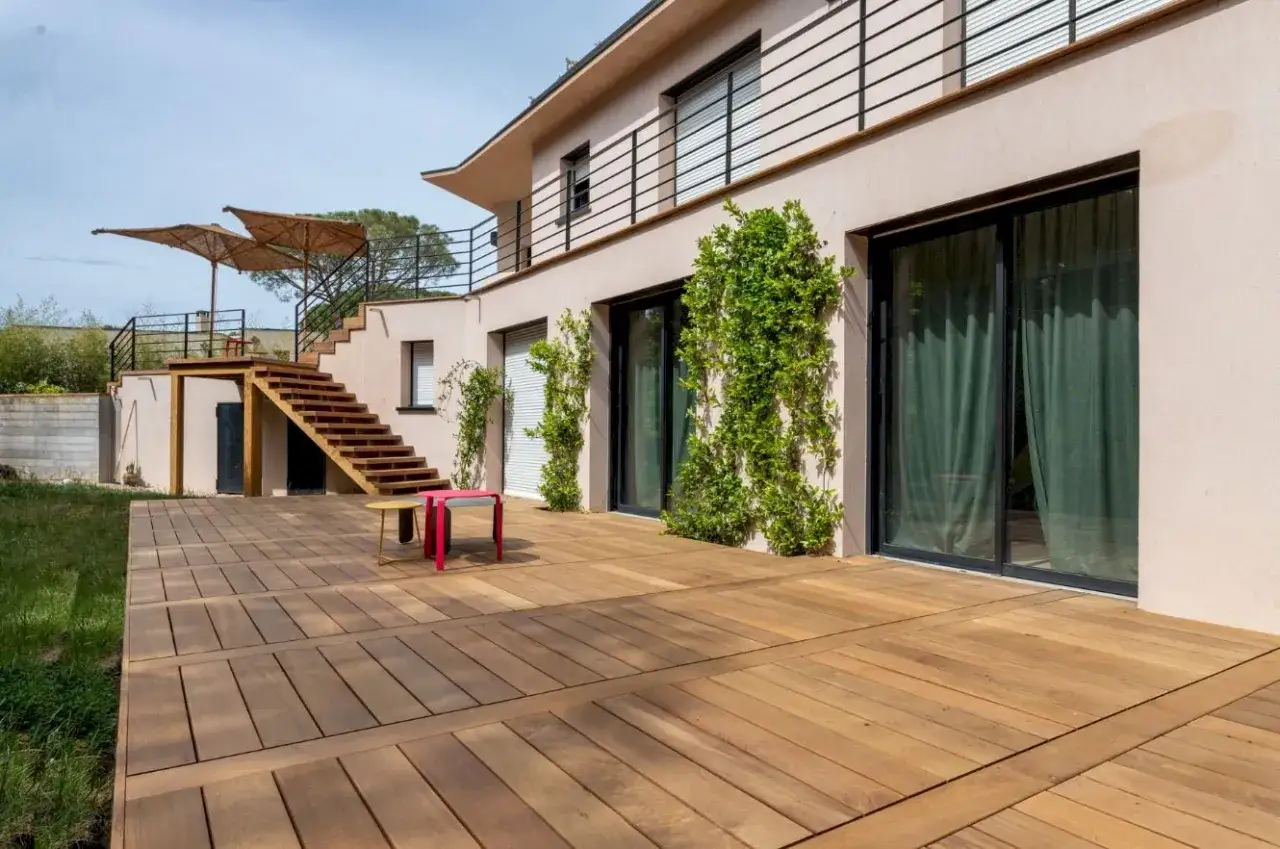 Une maison moderne avec **les plus belles terrasses en bois** s'ouvrant sur un jardin verdoyant. Des escaliers en bois mènent à un niveau supérieur avec des parasols.
