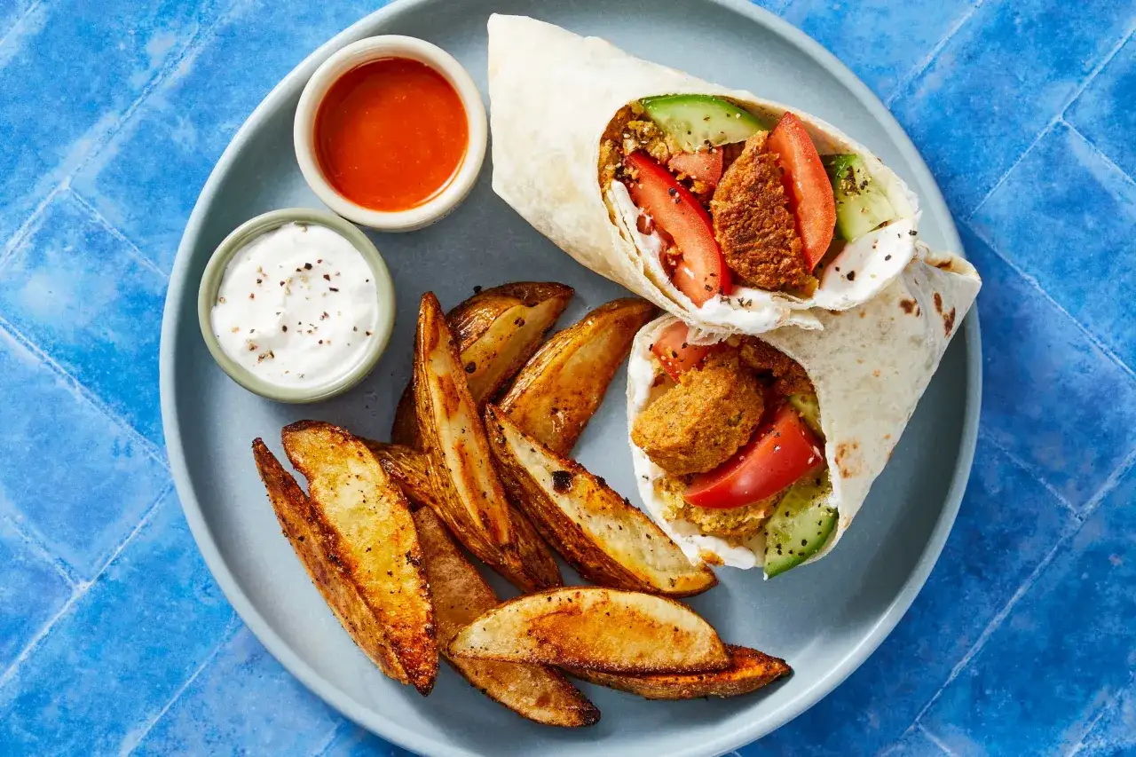 Två falafelwraps med tomat, gurka och vit sås, serveras med klyftpotatis och en chilisås. Ett perfekt wraps recept för en snabb middag.