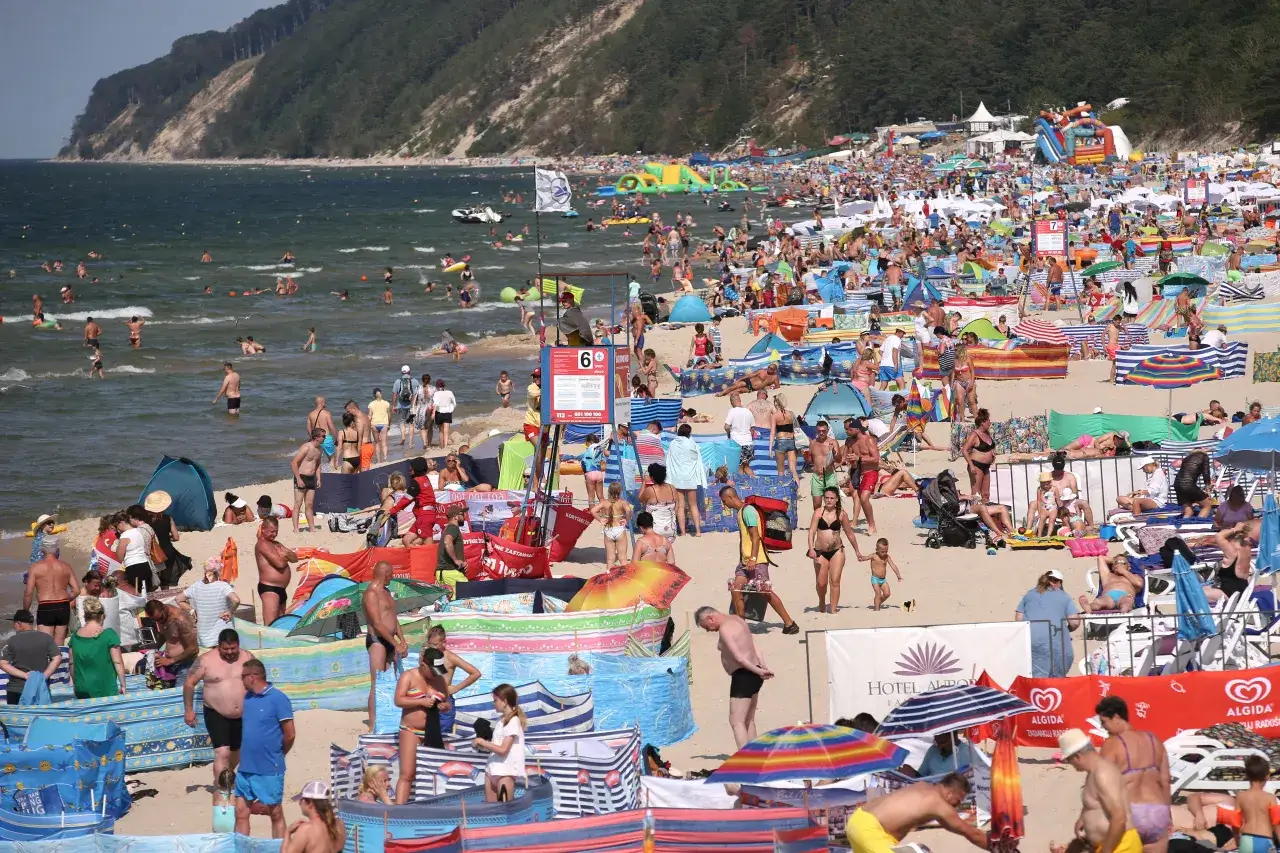 Tłum ludzi na plaży, w wodzie i na piasku. Wiele kolorowych parawanów i parasoli. W tle klif porośnięty lasem. Czy Morze Czarne jest ciepłe? Ludzie kąpią się i opalają, co sugeruje, że tak.
