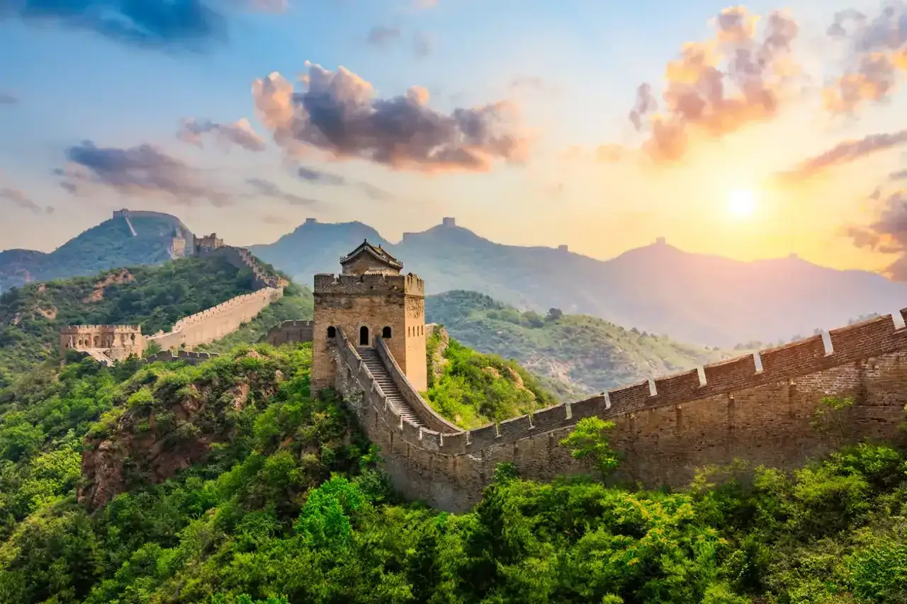 La muralla china serpentea entre montañas verdes al amanecer, con torres de vigilancia y un cielo anaranjado.