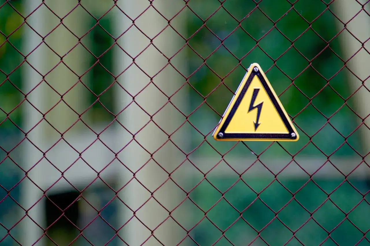 Warnschild an einem Maschendrahtzaun: Achtung, hier kann man leicht einen **Stromschlag bekommen**.