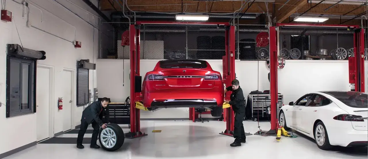 Mekaniker utf&ouml;r service p&aring; en r&ouml;d Tesla Model 3 som lyfts i en verkstad. En vit Tesla Model 3 st&aring;r bredvid.
