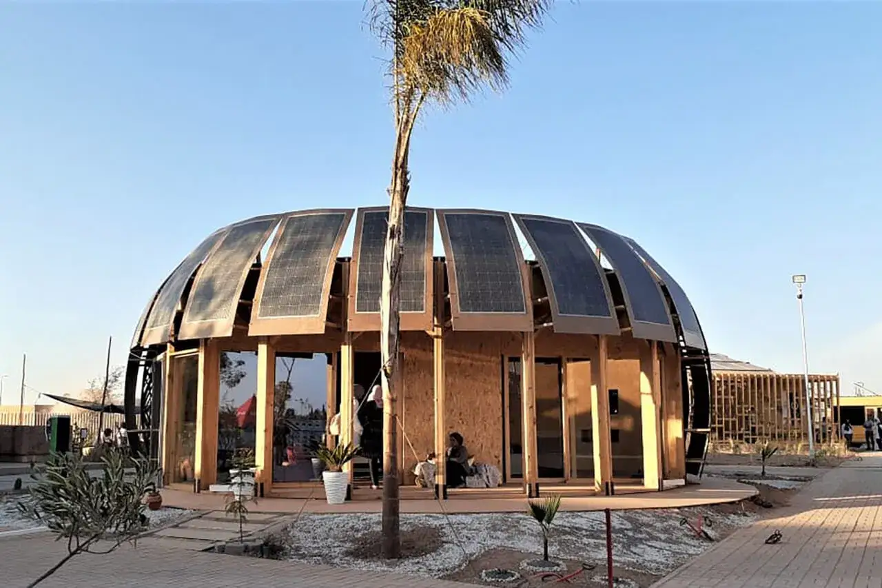 Edificio ecológico con paneles solares en su techo curvo, rodeado de vegetación y con un diseño moderno y abierto.