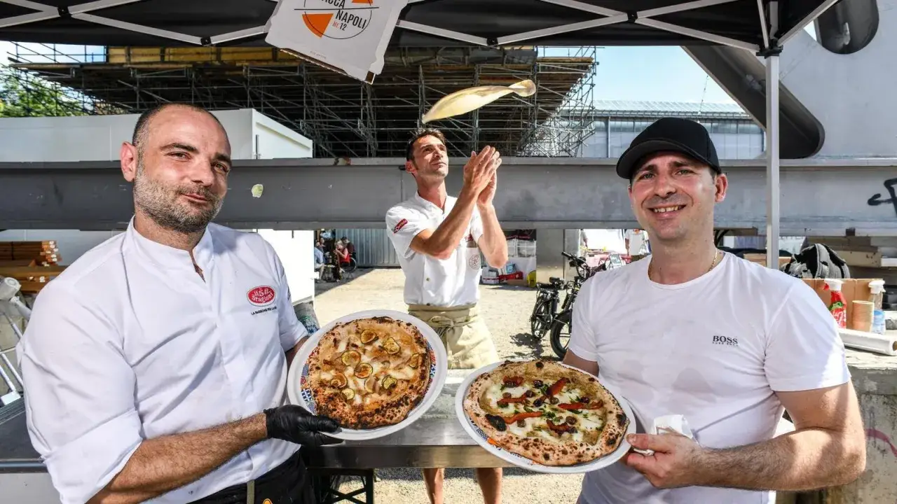 Drei Pizzabäcker präsentieren stolz ihre Kreationen. Einer wirft Teig in die Luft, während die anderen zwei fertige Pizzen zeigen. Ein Fest für die Sinne, das an