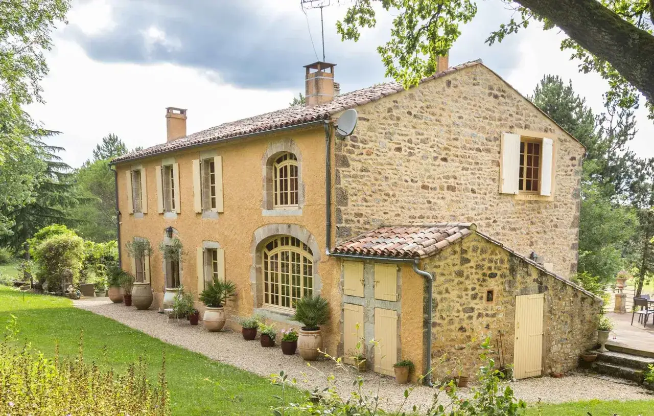 Belle maison de campagne avec un enduit &agrave; la chaux ocre, des murs en pierre et des volets jaunes. Un chemin de gravier serpente dans le jardin verdoyant.