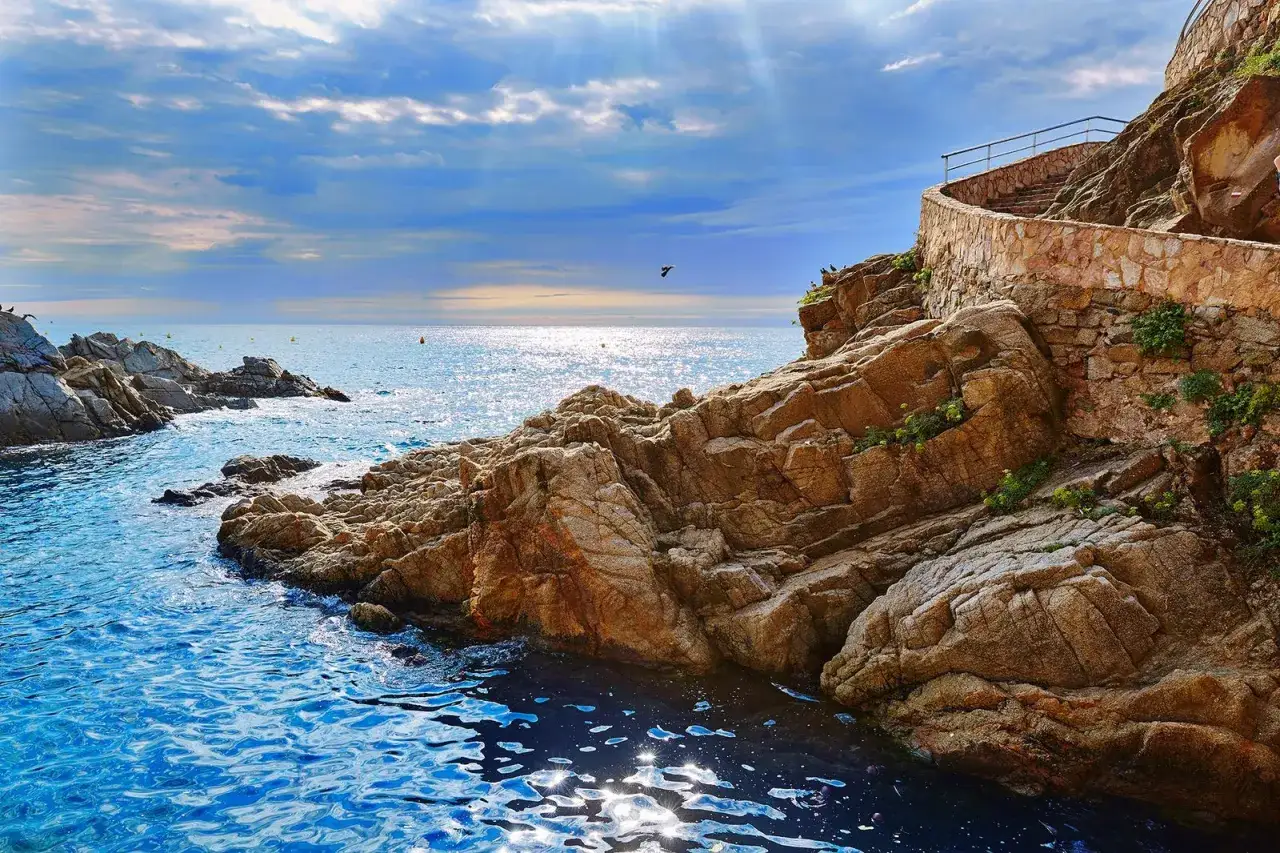 Escaleras de piedra ascienden por un acantilado rocoso junto al mar azul. Un **camino de ronda Costa Brava** invita a explorar la costa.