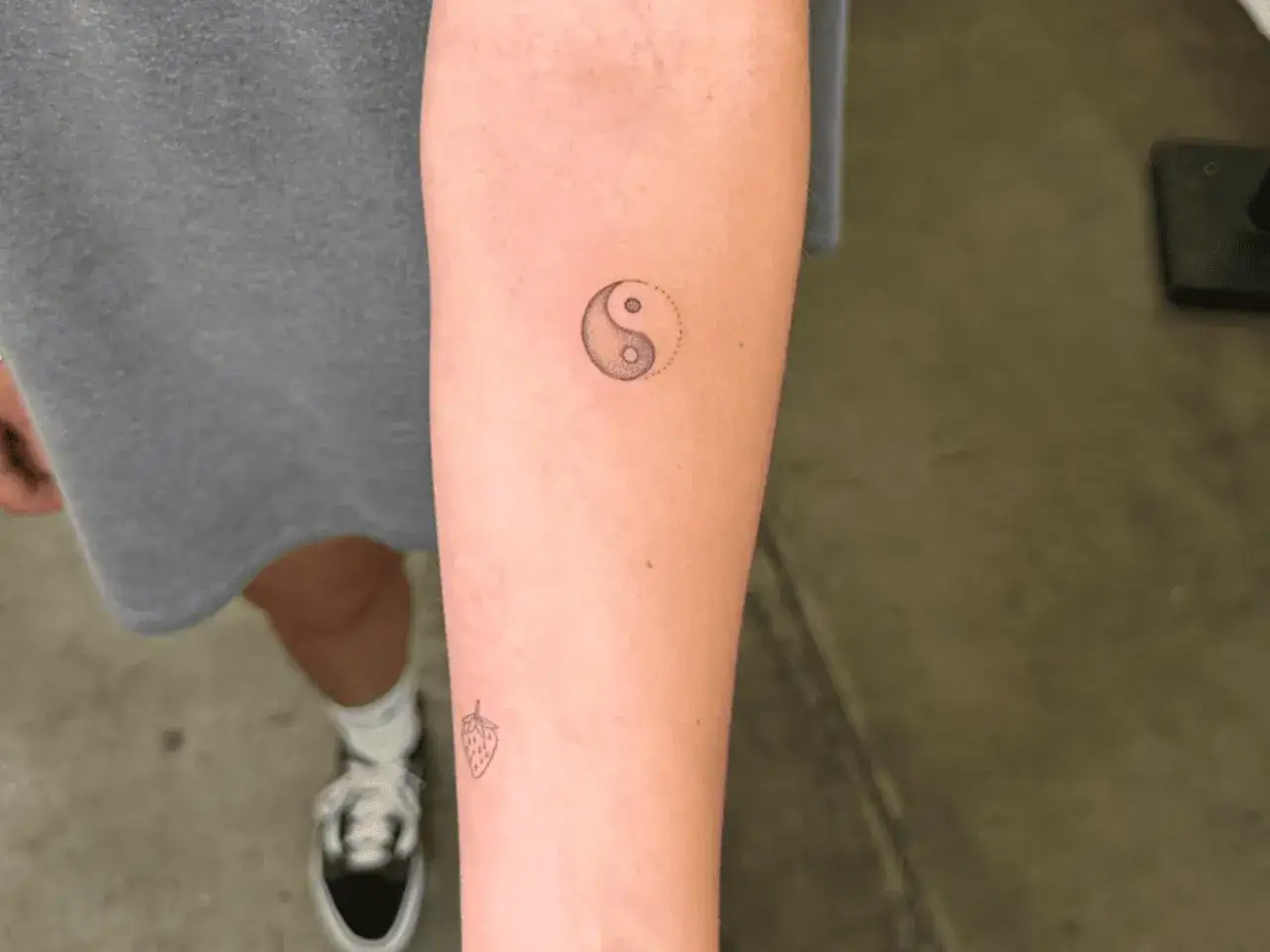 Delikatny tatuaż yin yang znaczenie r&oacute;wnowagi na przedramieniu, obok małej truskawki.