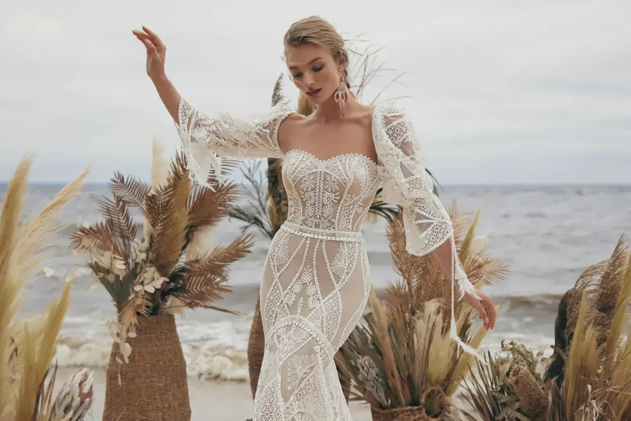 Piękna stylizacja boho na wesele. Koronkowa suknia z bufiastymi rękawami i ozdobnym paskiem, otoczona suchymi trawami i palmami na plaży.