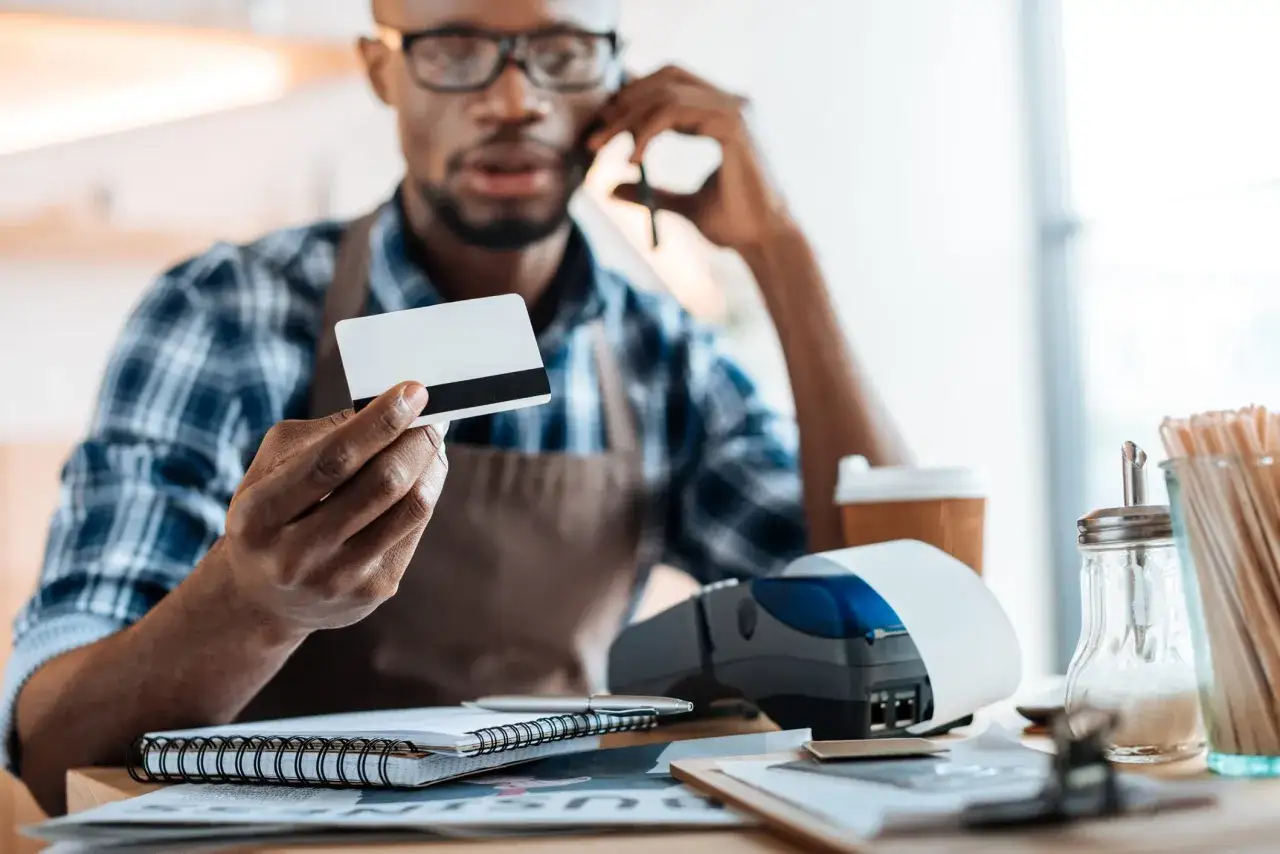 A man on the phone holds a credit card, pondering if you need an LLC for a business credit card while a payment terminal sits nearby.