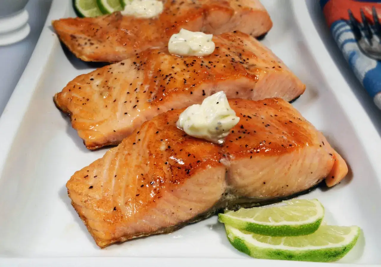 Drei saftige Flammlachs-Portionen mit einem Klecks Knoblauch-Dip und Limettenspalten auf einem weißen Teller.