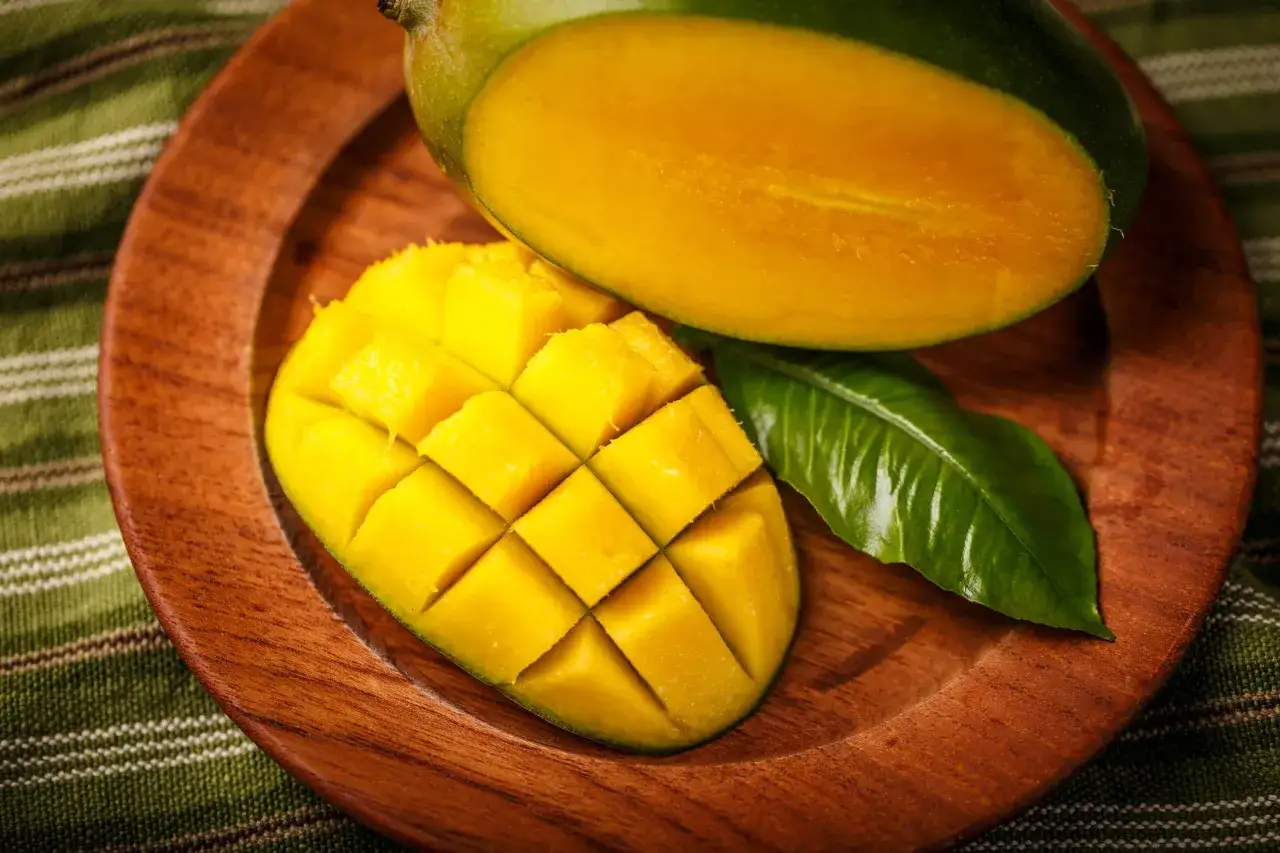 Soczysty mango, pełne witamin, pokrojone w kostkę na drewnianym talerzu. Obok leży połowa owocu i zielony listek.
