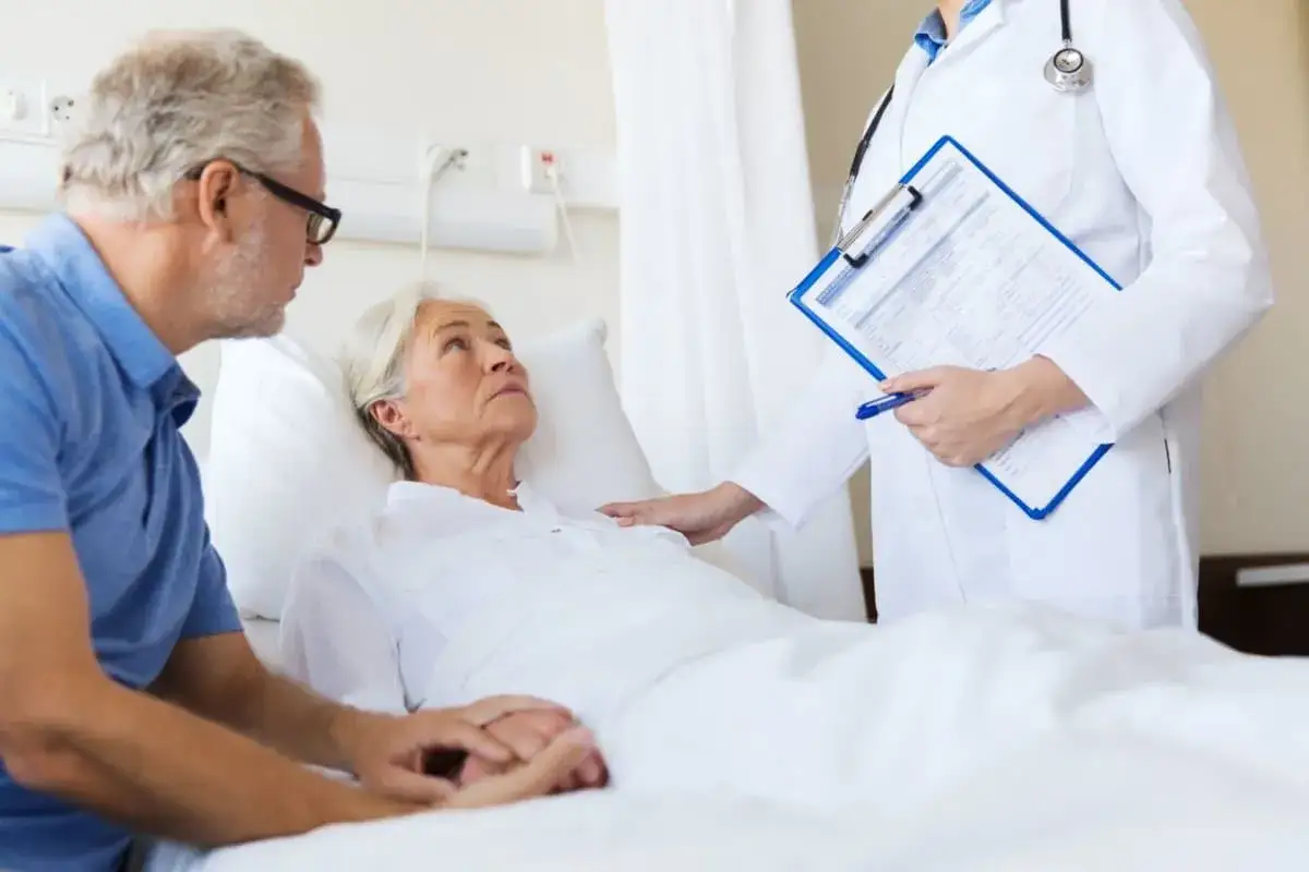 Älteres Paar im Krankenhaus. Frau liegt im Bett, Mann hält ihre Hand. Arzt mit Akte spricht über die **Chemotherapie abgelehnt prognose**.