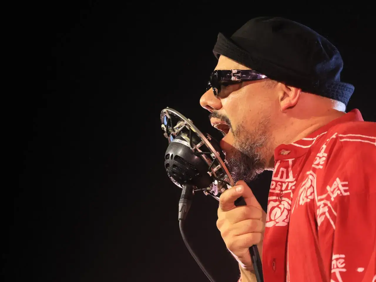 Un chanteur soul, barbe grisonnante, lunettes de soleil et bonnet noir, chante passionnément dans un micro vintage. Sa chemise rouge à motifs blancs est vibrante.