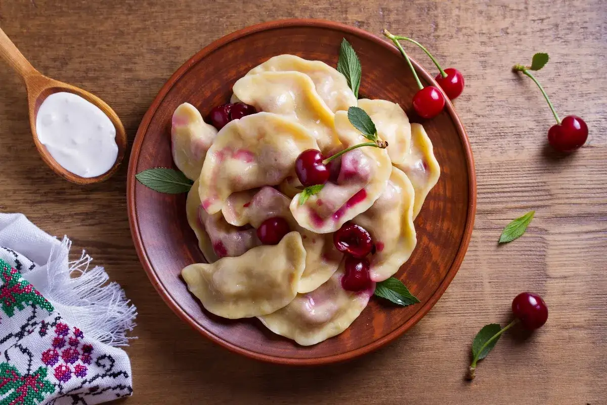 Pyszne pierogi z wiśniami na glinianym talerzu, obok łyżka śmietany. Idealne, gdy zastanawiasz się, jak odmrozić pierogi.