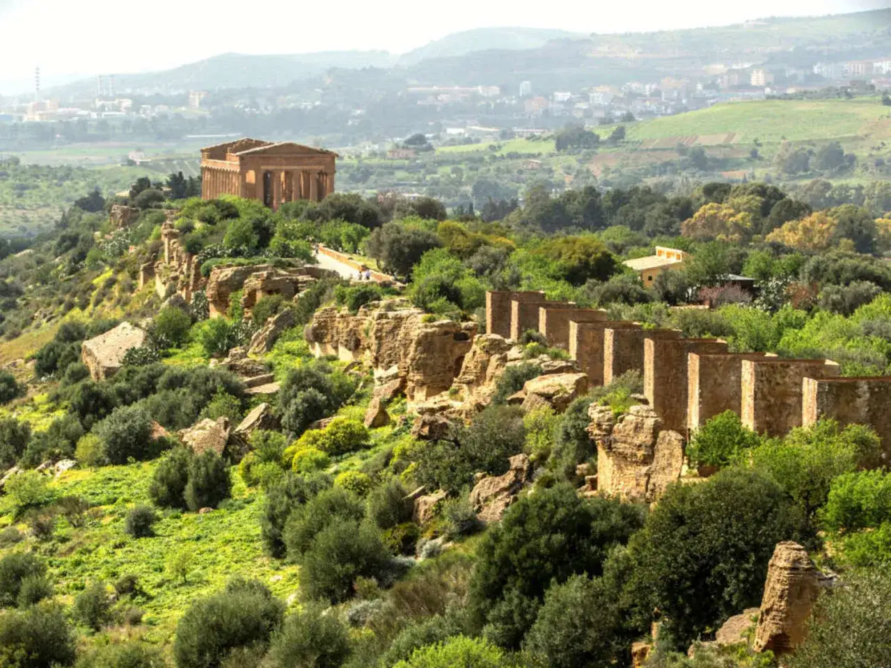 Ancient Greek temples stand majestically on a ridge in the Valley of the Temples, Sicily, surrounded by lush greenery and overlooking a distant town.