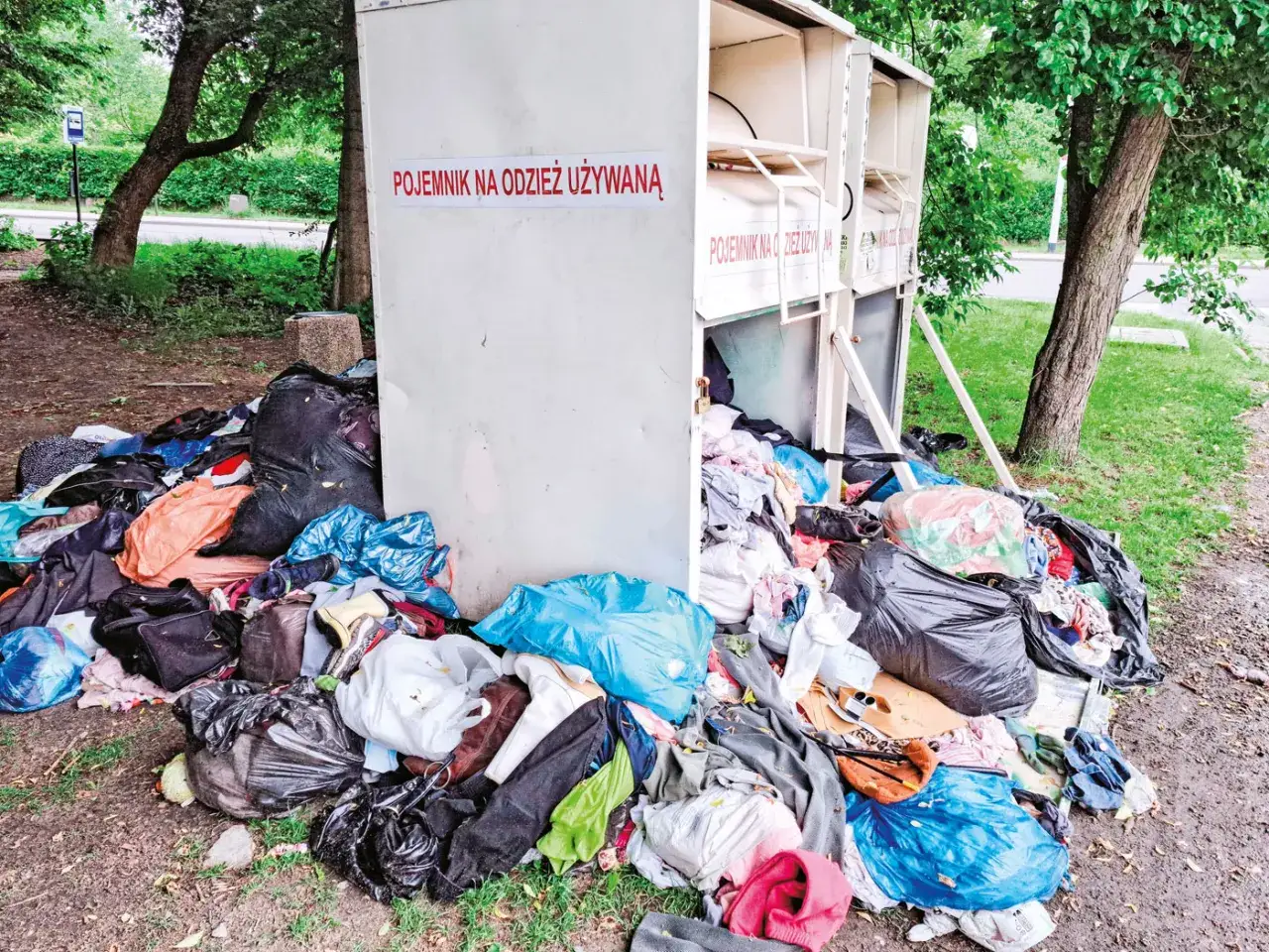 Kontenery na odzież używaną, otoczone stertami ubrań i worków. Czerwony krzyż na jednym z nich.