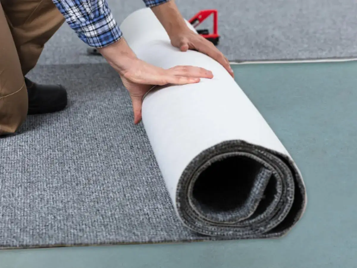 Mann rollt Teppich aus, um ihn zu verlegen. Grauer Teppichboden wird mit einer Rolle ausgelegt.