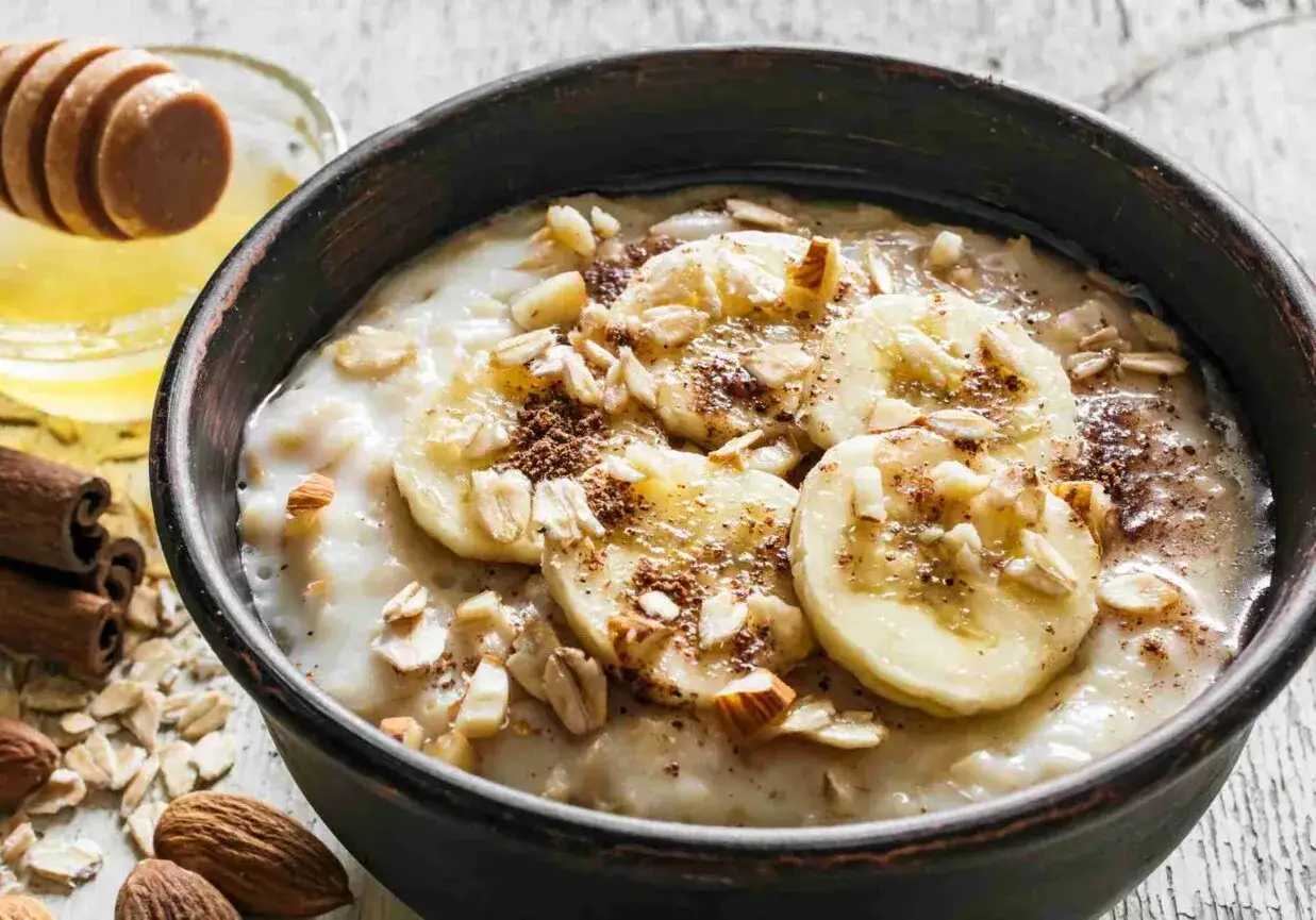 Pyszna owsianka z bananem, posypana płatkami migdałów i cynamonem. Obok miód i laskowe orzechy.