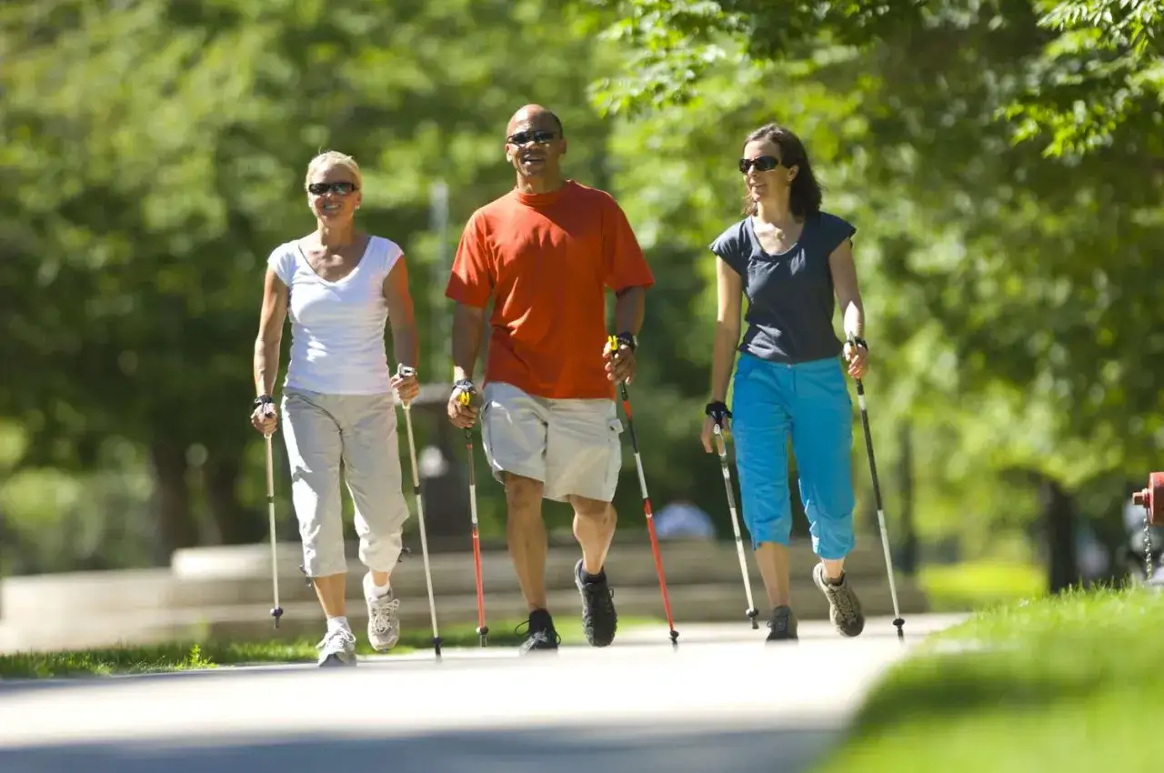 Trzy osoby w słoneczny dzień spacerują z kijkami. Pokazują, jak chodzić z kijkami, ciesząc się aktywnością na świeżym powietrzu.