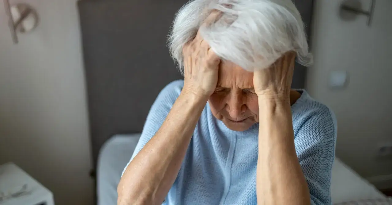 Starsza kobieta z rękami na skroniach, zamyślona. Może to być jeden z objawów alzheimera, trudność w skupieniu.