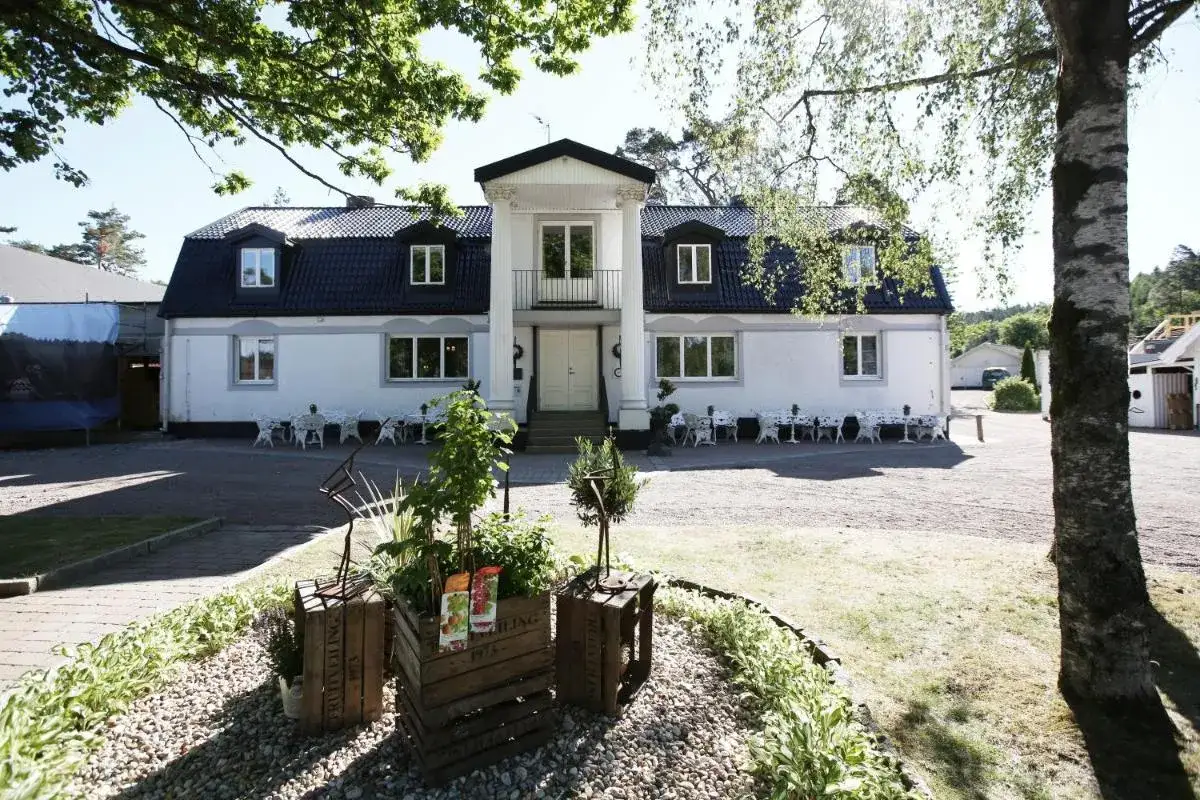 Vacker fasad p&aring; H&auml;llsn&auml;s Hotell & Restaurang med vita bord och stolar utomhus, omgivet av gr&ouml;nska.