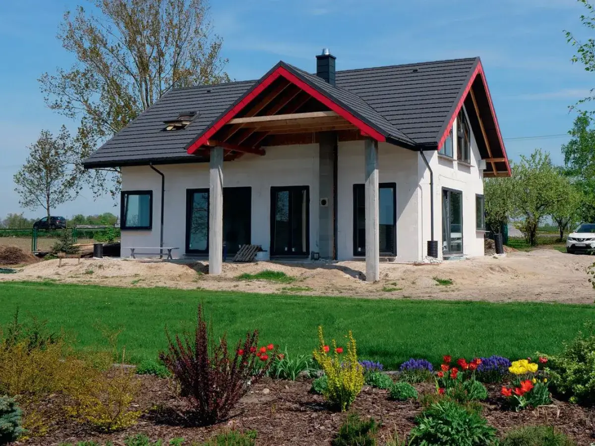 Nowy dom w stanie surowym, z czerwonymi akcentami dachu. Wiosna w pełni, kwitną kwiaty. Ciekawe, ile się czeka na odbiór domu.